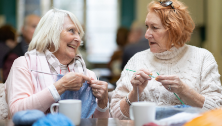 Two women in their 50s-60s chat together while knitting.